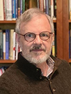 A gray haired white man wearing glasses sits in front of a bookshelf